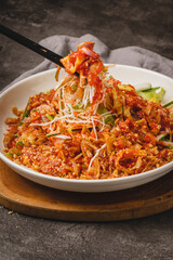 A close-up shot of a vibrant, spicy Asian noodle salad being served with chopsticks from a white bowl on a wooden board.