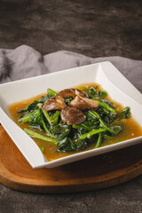 Stir-fried green leafy vegetables with shiitake mushrooms in a savory sauce served on a white square plate.