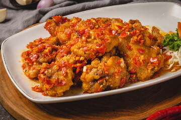 Crispy fried chicken pieces generously coated in a vibrant red chili sauce, served on a white plate with garlic and onions in the background.