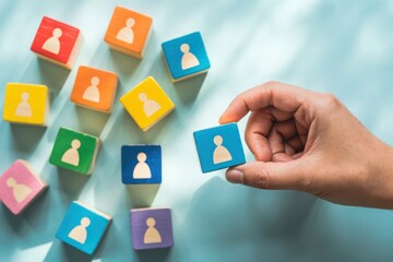 Hand selecting a blue wooden block with a person icon from a scattered group of colorful blocks on a light blue surface
