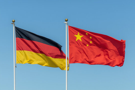 Flags of Germany and China waving next to each other on flagpoles against a clear blue sky, symbolizing bilateral relations and international diplomacy.
