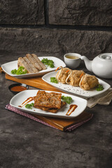 Three plates of various fried and savory Asian dishes, including sliced items, stuffed parcels, and fritters, served with a teapot and teacup on a dark textured surface.
