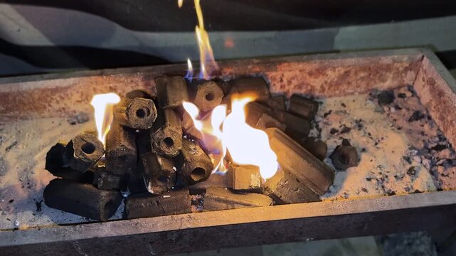 shot of glowing hexagonal charcoal briquettes burning on a bed of sand. Vibrant blue and orange flames dance in a rusty metal tray, creating intense heat and texture.