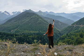 Naklejka premium A person stands on a rocky overlook, arms raised, gazing over distant mountains and lush landscape, capturing a moment of triumph in a vast outdoor scene.