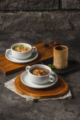 Two bowls of creamy mushroom soup garnished with croutons and herbs, served on wooden boards with a rustic wooden cup on a dark textured background.