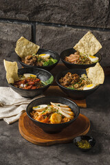 Five bowls of various Asian noodle dishes with different toppings and crackers, served on a wooden board on a dark textured surface.