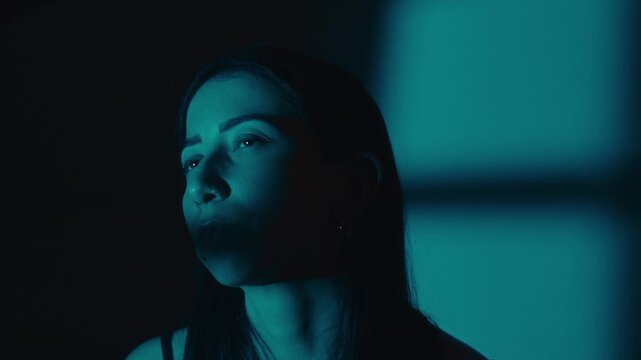 Woman looks up in teal lighting, with a square window shadow softly cast behind her on the wall.