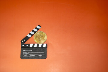 A Bitcoin coin rests on a film clapperboard against an orange background. This image suggests cryptocurrencies' potential in film production.