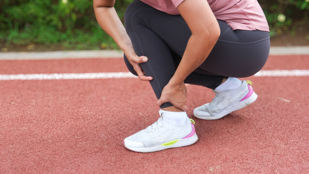 Woman runner holding leg suffering from muscle and tendon sprain pain in ankle at outdoor stadium, close-up. Athlete injured during intense workout. Health care and sport concept