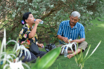An older couple sits barefoot and cross-legged on a manicured lawn, the woman drinks from a bottle while the man prepares a snack, enjoying a peaceful outdoor moment together.