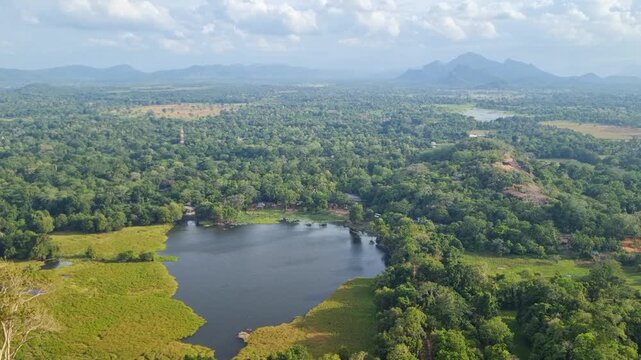 View from Lion Rock over the scenic heart of Sri Lanka, with a calm lake, lush greenery and forest unfolding beneath a partly cloudy sky, leading to distant mountains on the horizon
