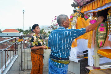 An older couple stands on an elevated balcony beside their decorated shrine, the man places incense while his wife holds yellow and white flowers, performing a daily ritual of devotion.