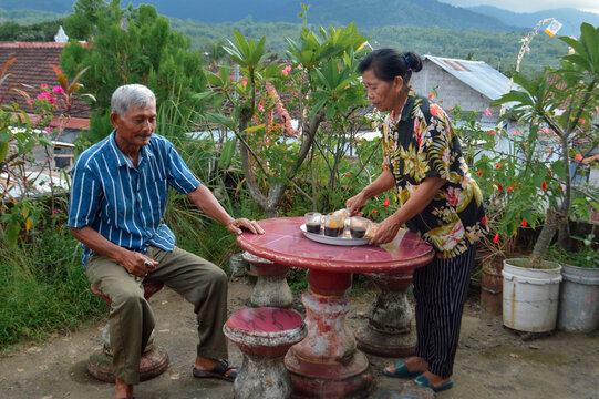 An older woman serves a tray with cups of coffee or tea and snacks to her husband, seated at a round red table with stone stools in a lush elevated garden overlooking village houses and mountains.