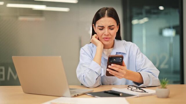 Woman sitting at office desk feels distressed, frustrated, and worried while intently viewing content on phone. Displays disappointment sadness, expressing strong emotional reaction to news received.