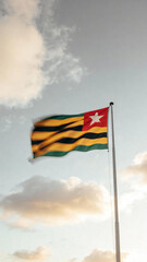 A flag with horizontal stripes of green and yellow, featuring a red square with a white star, waving against a backdrop of clouds and a soft sky.