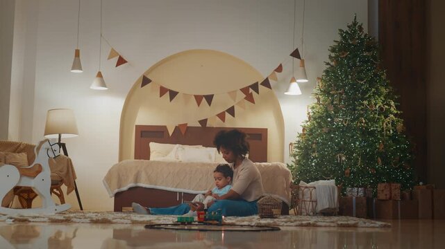 Beautiful decorated room in Christmas, mom and son playing toy railway on floor. African american woman and her little child watching cartoons and funny video for toddlers on smartphone screen