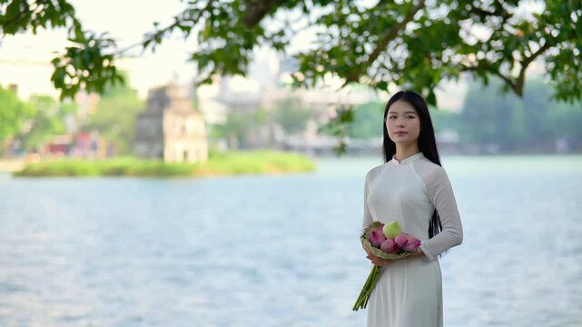 A young Vietnamese woman in a white ao dai standing by Hoan Kiem Lake, holding a bouquet of pink lotus flowers. Travel and Vietnamese culture