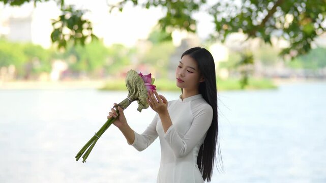 A young Vietnamese woman in a white ao dai standing by Hoan Kiem Lake, holding a bouquet of pink lotus flowers. Travel and Vietnamese culture