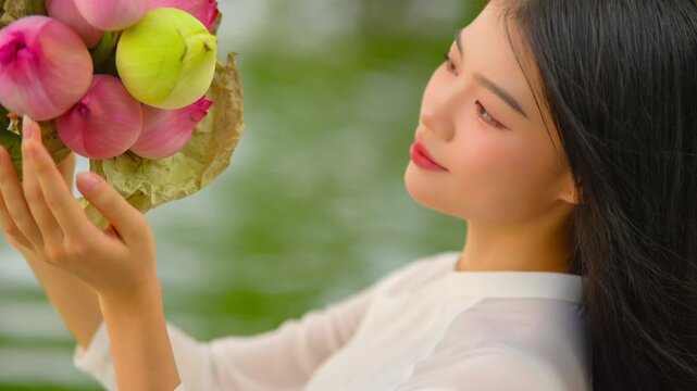 A young Vietnamese woman in a white ao dai sitting by Hoan Kiem Lake, holding a bouquet of pink lotus flowers