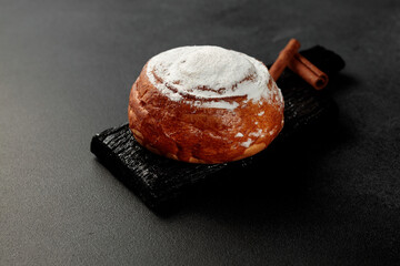 Round, delicious bun with cinnamon and powdered sugar on a black wooden board on a gray background