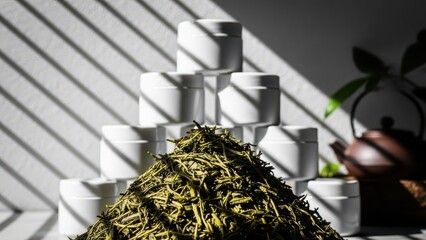 Sunlight casting dramatic striped shadows across a pyramid of white ceramic cups and a pile of dried green tea leaves