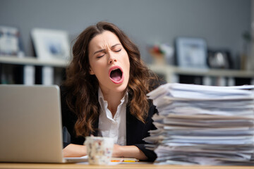 Lazy young employee woman yawning and bored with stack of paperwork or document folder on desk in office.