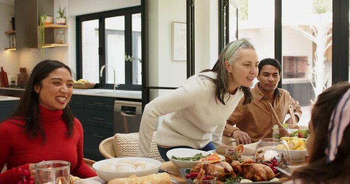 Mature woman standing at home table and thanking guests, sparking laughter, family serving meal