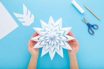 Young adult woman hands holding and showing white snowflake shape from paper on blue table background. Pastel color. Making decoration element for winter festive. Closeup. Flat lay. Top down view.
