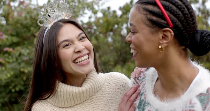 Diverse female friends exchanging eye contact and laughing wearing tiara and red headband in garden