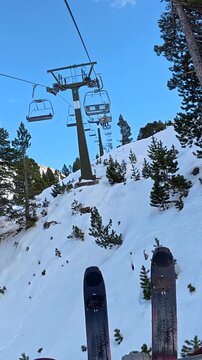 Personal perspective of a skier riding a chairlift up a snow covered mountain, with skis visible in the foreground and a scenic alpine landscape below on a bright, sunny winter day