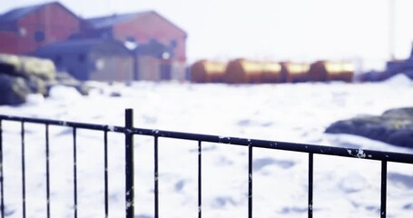 A snowy landscape shows a metal fence in the foreground, with industrial buildings and storage tanks partially obscured by falling snow in the background, creating a serene winter scene. - Powered by Adobe