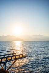 Obraz premium A peaceful sunset scene featuring a wooden pier overlooking calm ocean waters, with golden sunlight reflecting across the sea. Soft mountain silhouettes fade into the warm evening haze