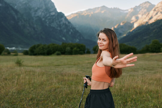 A cheerful woman in athletic wear stands in a wide meadow, distant mountains in view, extending her arm and smiling, capturing outdoor energy and a sense of adventure. - Powered by Adobe