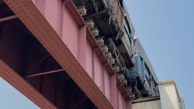 Freight wagons rumble across a steel railway bridge, captured from a low static angle showing metal beams, wheels, and vibrant industrial details.