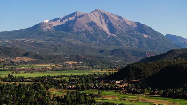 Lush valley spreads below Mount Sopris with rich greens contrasting the rugged Colorado summit, aerial tilt up establishing
