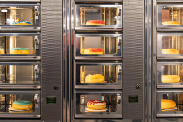 Automated vending machine filled with various colorful glazed donuts behind glass doors, offering sweet treats for sale in a public shopping area.