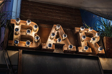 Vintage rusty metal BAR sign with light bulbs and spiderwebs, mounted on a wall and illuminated in a dim urban setting with plants in the background.