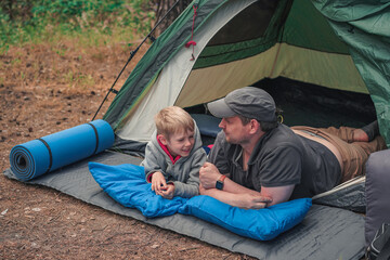 Father and smiling son lie on their stomachs talking near a tent entrance, sharing a sweet moment at a forest campsite.