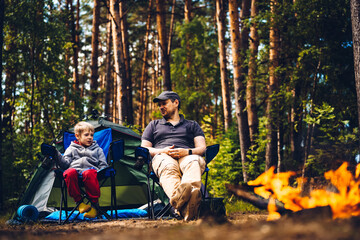 Father and son sit side-by-side in chairs by a bright campfire, relaxing in front of a tent at a shady forest campsite.