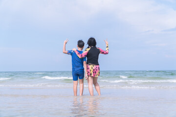 Asian mother and son hugging by the sea and raising peace signs, back view
