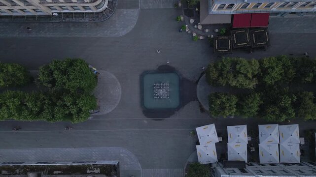 An aerial view of a public square with a fountain, trees, and cafe umbrellas. A few people and cyclists are present.