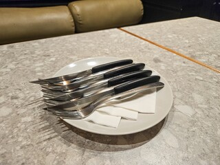 A stack of clean forks and knives with black handles resting on a white plate with napkins on a textured table.