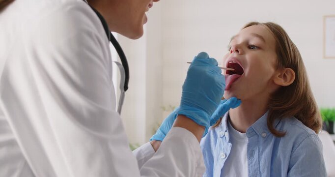 Child pediatrician doctor, otorhinolaryngologist examining little boy throat during visit in clinic, holding wooden spatula tongue reviewing patient inflamed tonsils, provide professional medical aid