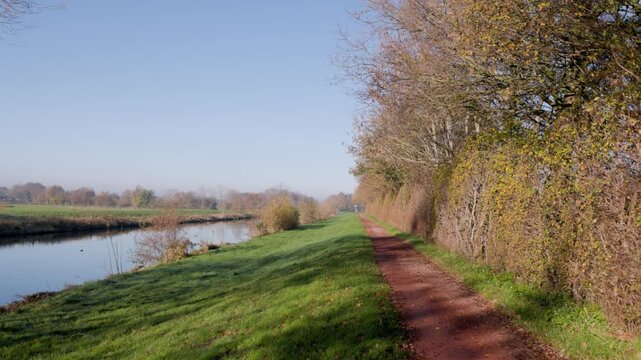 Dirt path beside calm river bordered by leafless trees and grassy bank clear sky and soft light evoke peaceful late autumn atmosphere.
