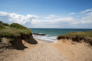 sandy path through grassy dunes toward a peaceful shoreline, gentle waves, beach, bright blue sky, scattered clouds. Coastal landscape, calm, inviting seaside atmosphere, travel, nature, relaxation