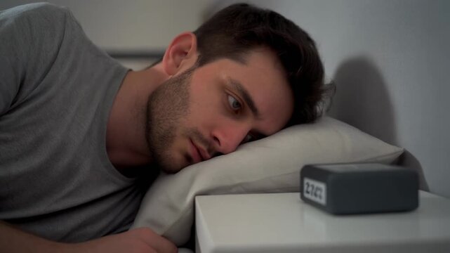 Tired man in bed hitting the snooze button on his alarm clock, struggling to wake up in the morning in his bedroom