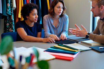 Young adult Caucasian man explaining ideas to young adult Black woman and young adult Caucasian woman during business meeting at table with fabric samples and notebooks