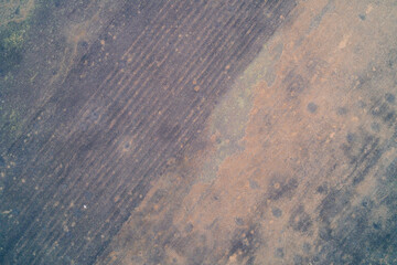 Abstract top-down aerial view of textured field with diagonal tillage lines and rust colored soil patches. Rough rural ground background.