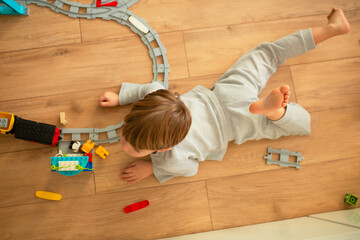 
Вид сверху: ребёнок лежит на полу и играет с игрушечной железной дорогой. Тёплая домашняя атмосфера, концепт детского творчества и раннего развития.