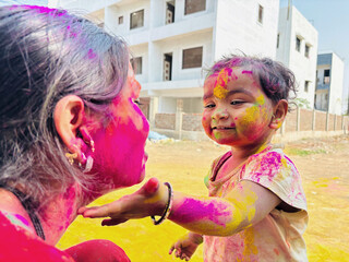 An Indian mother and child enjoying Holi as the child gently applies bright colors to her face, both smiling and covered in vibrant powder while standing outdoors near residential buildings in warm da